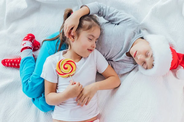 Encantadora hermana está durmiendo sobre el vientre de su hermano, sosteniendo una gran piruleta en un palo, ambos se ven dulces . — Foto de Stock