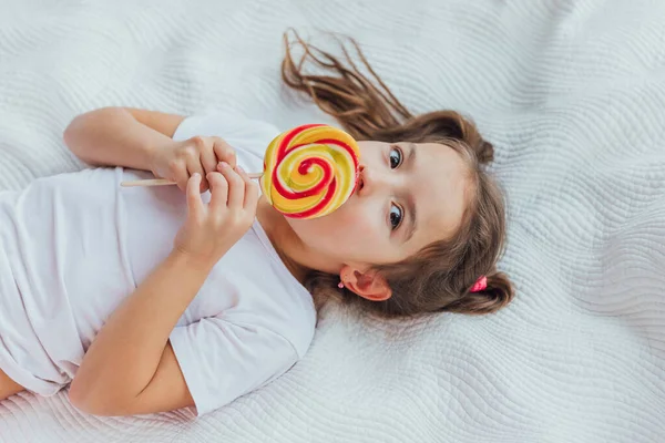 Gorgeous kid with funny pigtails is licking red-yellow candy on stick, smiling lovely to the camera. — Stock Photo, Image