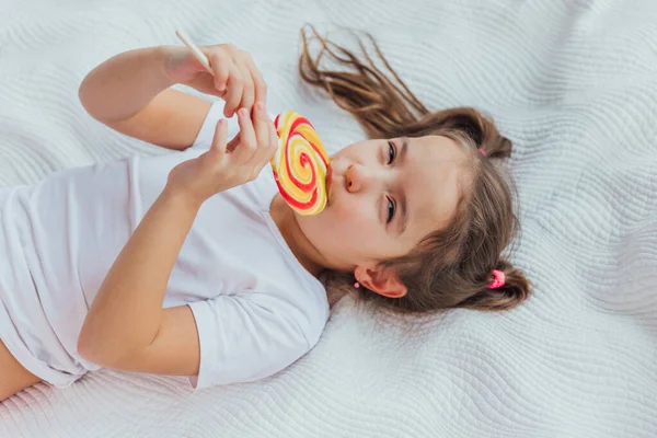 Hermoso niño con coletas divertidas está lamiendo caramelos rojo-amarillo en palo, sonriendo encantadora a la cámara . —  Fotos de Stock