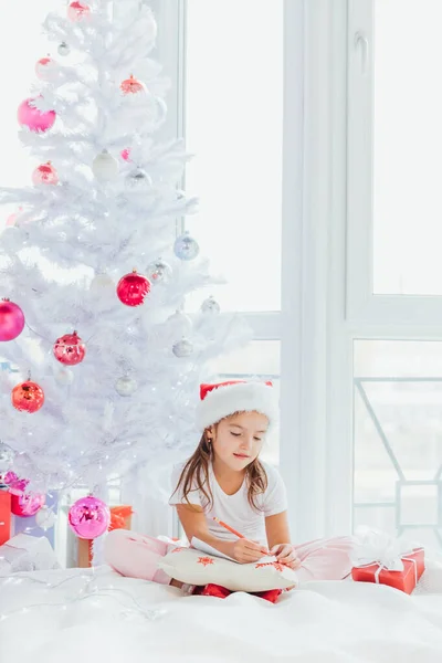 Bambina in cappello Babbo Natale scrive lettera a Babbo Natale vicino all'albero di Natale nella fredda mattina d'inverno . — Foto Stock