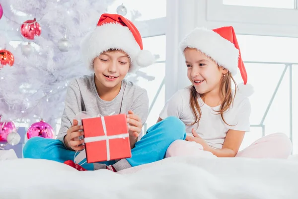 Δύο παιδιά που φορούν καπέλα santa και πιτζάμες και μοιράζονται κουτιά χριστουγεννιάτικων δώρων σε Χριστουγεννιάτικη ατμόσφαιρα. — Φωτογραφία Αρχείου