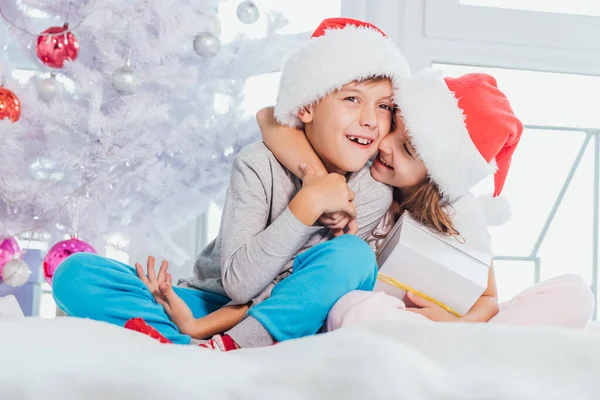 Due bambini che indossano cappelli e pigiami di Babbo Natale e condividono scatole regalo di Natale in atmosfera natalizia progettata . — Foto Stock