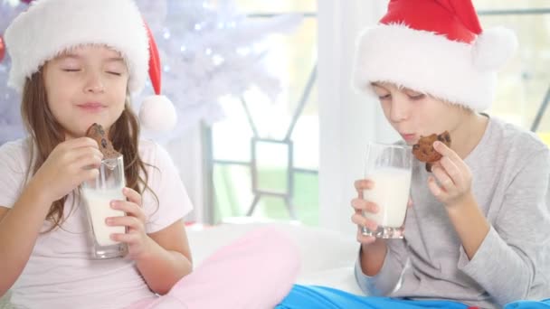 Carino i bambini piccoli a Babbo Natale cappelli bere latte e mangiare deliziosi biscotti a casa, cercando pienamente soddisfatto . — Video Stock