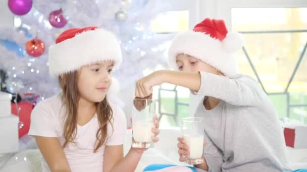 Cute little kids in Santa kapelusze picia mleka i jedzenia pyszne ciasteczka w domu, patrząc w pełni zadowolony. — Wideo stockowe