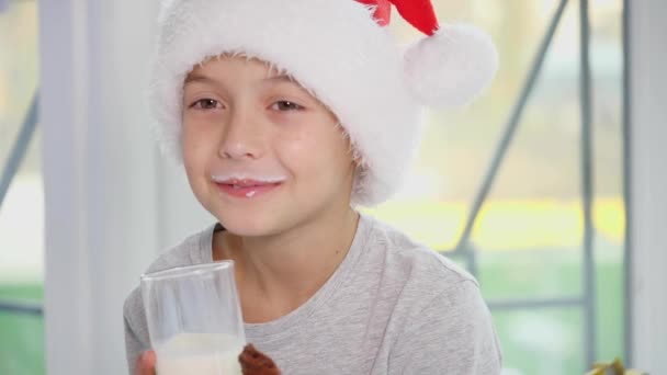 Zugeschnittenes 4k-Video eines kleinen Jungen im Weihnachtsmannhut, der Milch trinkt und leckere Kekse isst, Milch leckt und Schnurrbart trägt. — Stockvideo