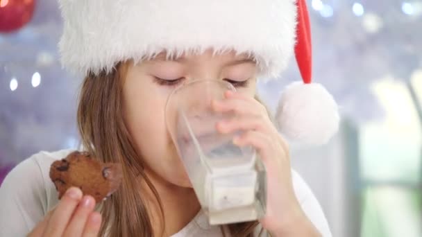 Ritagliato 4k video di bambina a Babbo Natale cappello bere latte e mangiare deliziosi biscotti a casa . — Video Stock