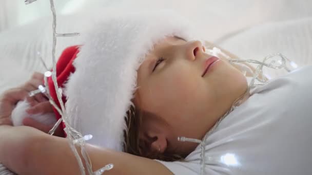 Κοντινό βίντεο του χαριτωμένου μωρού σε santa καπάκι, που βρίσκεται στο κρεβάτι, παίζει με γιρλάντες κρέμονται από το κλαδί έλατο, χαμογελώντας υπέροχο. — Αρχείο Βίντεο