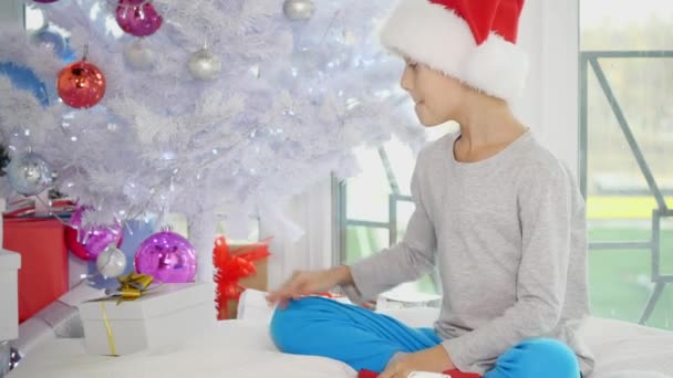 有趣的视频：好奇的男孩从树下拿圣诞礼物，摇晃着，敲着，听着，想知道里面装的是什么. — 图库视频影像