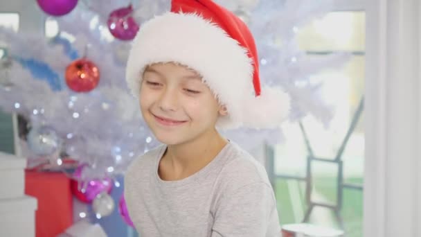 4k video Santa chlapec sedí u vánočního stromku, hraje si s cukrovinkami, dívá se přes cukrovou třtinu srdce, uvedení jazyk ven, usmívá. — Stock video