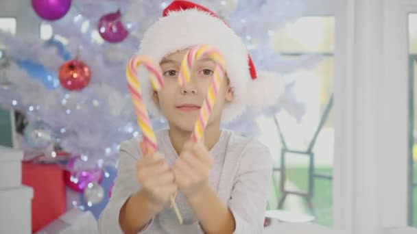 Video of funny boy in santa hat sitting on the bed in pyjamas, extending striped candy canes to the camera, winking. — Stock Video