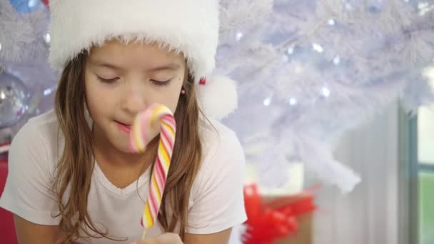 Pretty little girl is twisting in hands a candycane, licking it sometimes, looking curious. — Stock Video