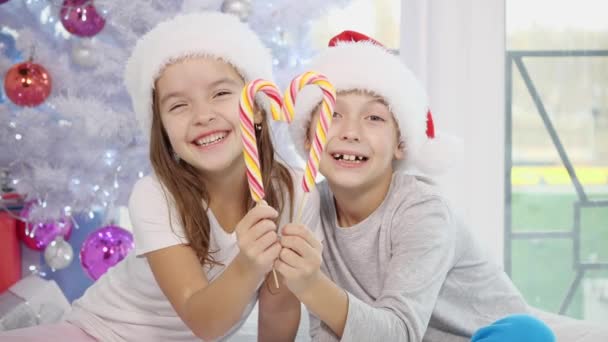 Adorables niños caucásicos disfrutan de la mañana de Navidad, jugando en la cama, haciendo corazón con bastones de caramelo, luego lamiendo caramelos a rayas desesperadamente . — Vídeo de stock