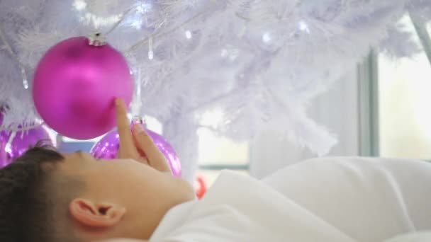 Vídeo recortado de un niño pequeño acostado bajo el árbol de Navidad, jugando con bolas de color rosa . — Vídeo de stock