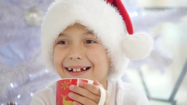 Vídeo recortado de hermoso niño en sombrero de santa, cerca del árbol de Navidad, con una taza de cacao, mirando encantado y complacido sobre fondo blanco brillante . — Vídeo de stock