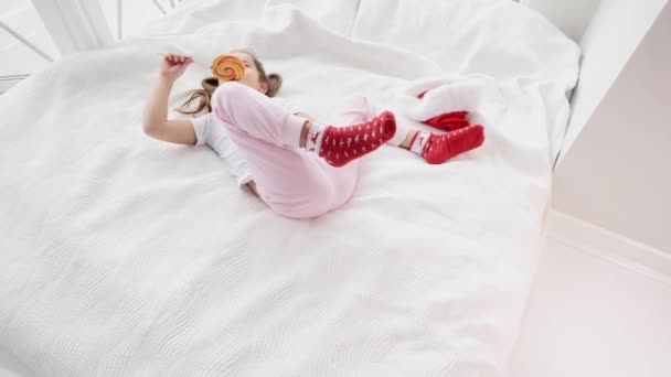 Dulce vídeo de precioso niño con divertidas coletas despertando y lamiendo caramelo rojo-amarillo en palo, disfrutando del momento . — Vídeos de Stock