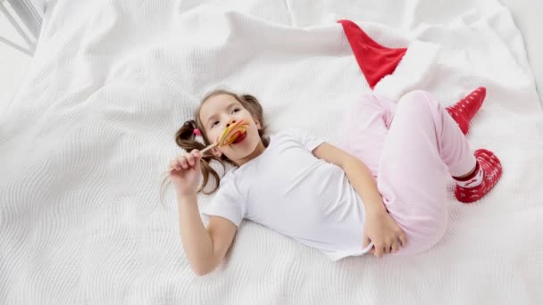 Sweet vídeo of gorgeous kid with funny pigtails wake up and licking red-yellow candy on stick, aproveitando o momento . — Vídeo de Stock