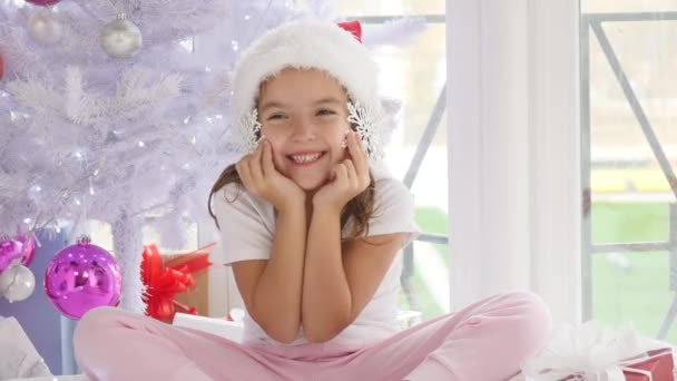 Pequeña princesa sentada cerca del mágico árbol de navidad y sosteniendo copos de nieve decorativos como si fueran auriculares o pendientes y mirando a la cámara con la expresión facial más dulce de la historia . — Vídeo de stock
