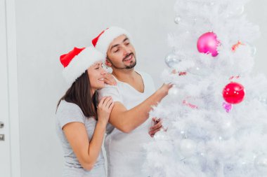 Mutlu çift Noel ağacını süslüyor. Sakallı adam ve yeni yıl şapkalı esmer kadın evde Noel ağacı süsleyerek eğleniyorlar.