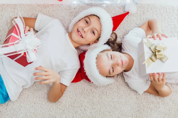 男孩和女孩躺在圣诞树附近的地板上, 带着礼物。冬天家里戴红帽子的孩子们 — 图库照片