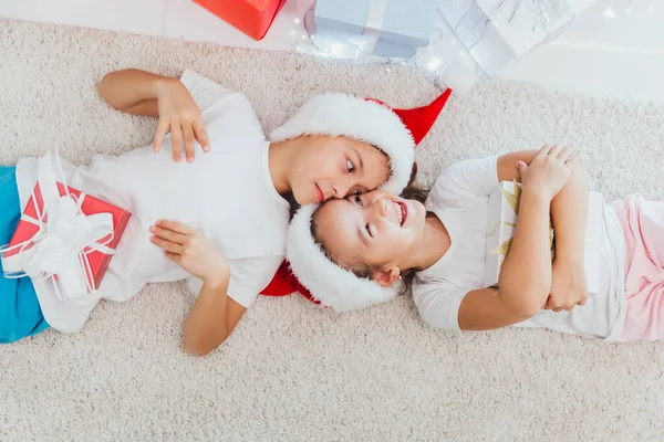 Хлопчик і дівчинка лежать на підлозі з подарунками біля ялинки. Діти в червоних капелюхах вдома взимку — стокове фото