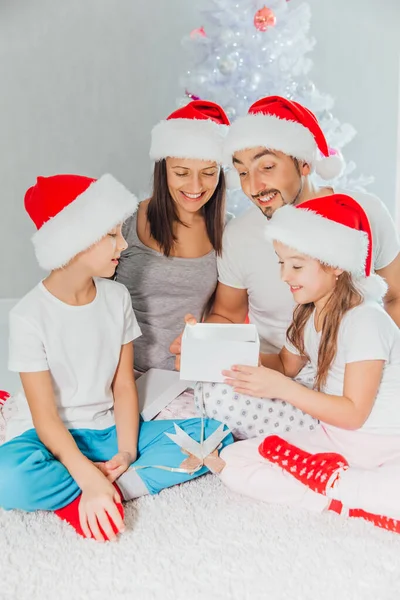 Young mother, father and two little children opening a magical Christmas gift by a Christmas tree in cozy living room in winter — Stock Photo, Image