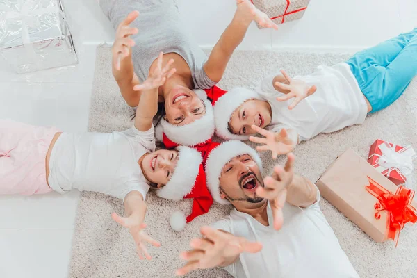 Красивая молодая семья с маленькими детьми лежит у новогодней елки с подарками на праздник — стоковое фото