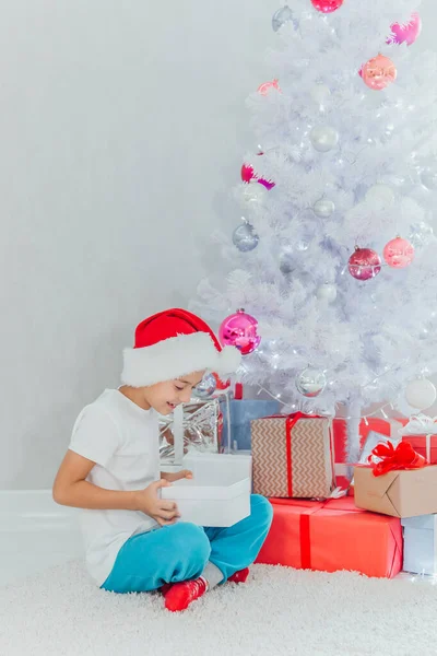 Αγόρι με καπέλο Άι Βασίλη που κρατάει δώρα κοντά στο χριστουγεννιάτικο δέντρο. Χριστουγεννιάτικη διακόσμηση. Πρωτοχρονιά και Χριστούγεννα. Μαγικές στιγμές. Εσωτερική φωτογραφία — Φωτογραφία Αρχείου
