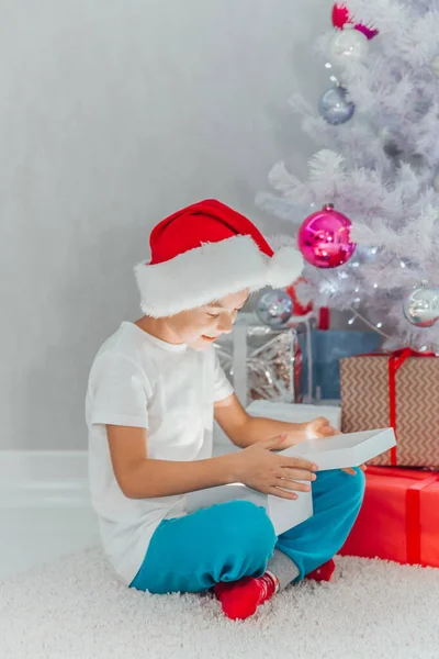 Feliz niño sonriente en sombrero de santa cerca del árbol de Navidad con regalo. Decoración de Navidad. Concepto de Año Nuevo y Navidad. Tiempo mágico. Foto interior — Foto de Stock