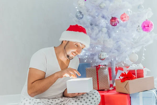 Młody człowiek w kapeluszu Mikołaja siedzi pod choinką i otwiera pudełko prezentów przed białymi światłami wczesnym rankiem — Zdjęcie stockowe