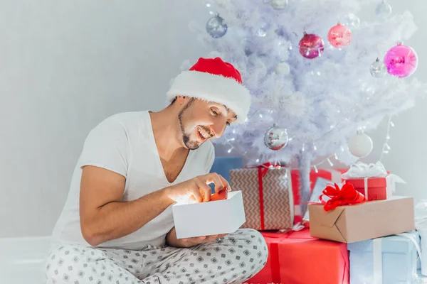 Νεαρός άνδρας με τα Χριστούγεννα κοστούμι άνοιγμα λαμπερό δώρο, κάθεται δίπλα στο χριστουγεννιάτικο δέντρο — Φωτογραφία Αρχείου
