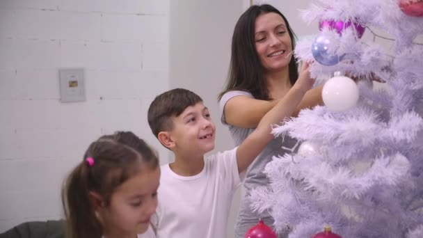 Η μητέρα και τα δύο παιδιά στολίζουν ένα χριστουγεννιάτικο δέντρο με πολύχρωμους θάμνους στο σαλόνι.. — Αρχείο Βίντεο
