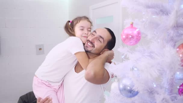 Hermosa niña pequeña abrazando a su padre, abrazando firmemente su cuello en la casa cerca del abeto . — Vídeos de Stock