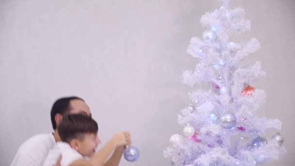 El pequeño hijo quiere colgar una bola rosa en la rama superior del árbol, así que el padre lo recoge, el niño hace lo que quiere y papá muestra la lengua a la cámara . — Vídeos de Stock