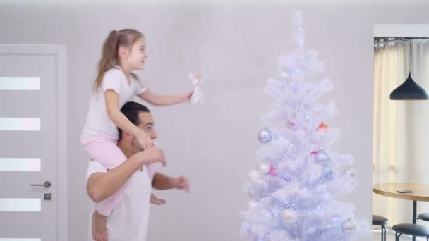 Vater und Tochter sitzen auf seinem Hals, kommen zum silbernen Weihnachtsbaum und Tochter setzt Engelsschmuck in die Baumkrone. — Stockvideo