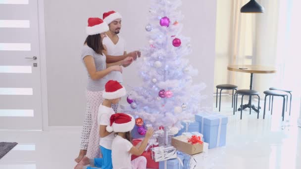 Feliz familia hablando riendo y decorando juntos el árbol de navidad, vestidos con pijamas y sombreros de santa . — Vídeo de stock