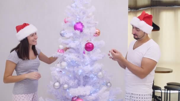 Divertida esposa y marido de pie en diferentes lados del árbol de navidad de plata, tonteando y decorando, vestido con bonitos sombreros de santa . — Vídeo de stock