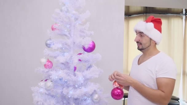 Hombre barbudo divertido en sombrero de santa está quitando bolas del árbol de christmas . — Vídeos de Stock