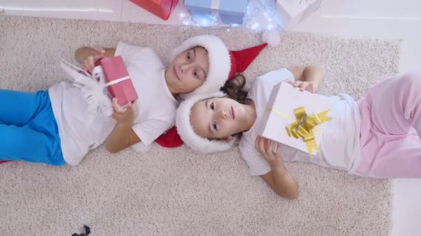 Top view video of cute children lying on the white carpet, shaking, knocking, listening to what is inside Christmas presents. — Stock Video