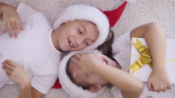 Dos niños con sombreros y pijamas de santa, tumbados cabeza a cabeza en la alfombra con cajas de regalo de Navidad en el vientre y haciendo muecas tontas, mirando a la cámara . — Vídeo de stock