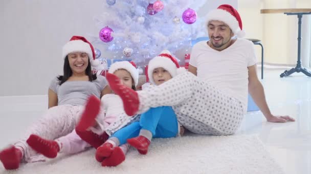 Joyeux père mère soeur frère sont prêts pour Noël, assis sur le tapis près de sapin, vêtus de chapeaux de Père Noël et chaussettes rouges, jouer autour . — Video