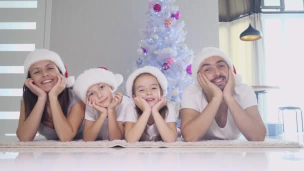 Divertido padre madre e hijos en calcetines rojos y sombreros están acostados en la alfombra cerca del árbol de Navidad, manteniendo las manos en las mejillas y moviendo la cabeza de lado a lado . — Vídeos de Stock