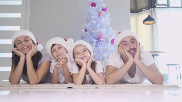 Vtipný otec matka a děti v červených ponožkách a kloboucích leží na koberci u vánočního stromku, drží ruce na tvářích a pohybuje hlavou ze strany na stranu. — Stock video