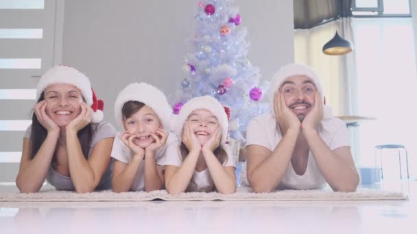 Funny father mother and children in red socks and hats are lying on the carpet near christmas tree, keeping hands on cheeks and moving head from side to side. — Stock Video