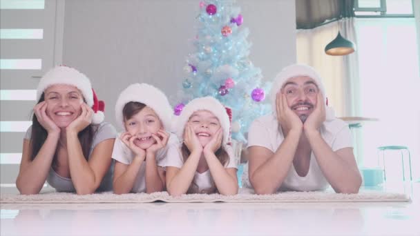 Grappige vader moeder en kinderen in rode sokken en hoeden liggen op het tapijt bij de kerstboom, houden handen op de wangen en bewegen van links naar rechts. — Stockvideo