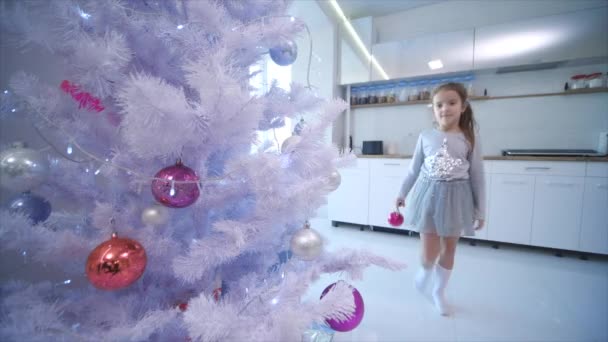 Menina morena pequena está decorando uma árvore de Natal falsa com bugigangas coloridas em casa . — Vídeo de Stock
