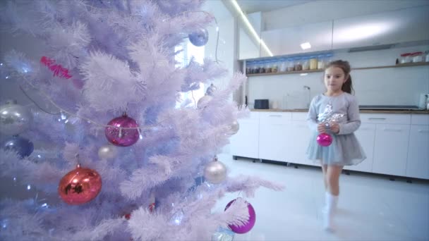 Vidéo 4k de petite fille brune accrochant une boule de verre rose sur un faux sapin de Noël à la maison . — Video