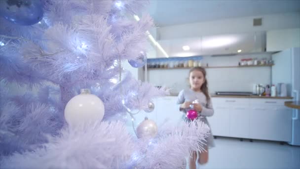 Vidéo 4k de petite fille brune accrochant une boule de verre rose sur un faux sapin de Noël à la maison . — Video