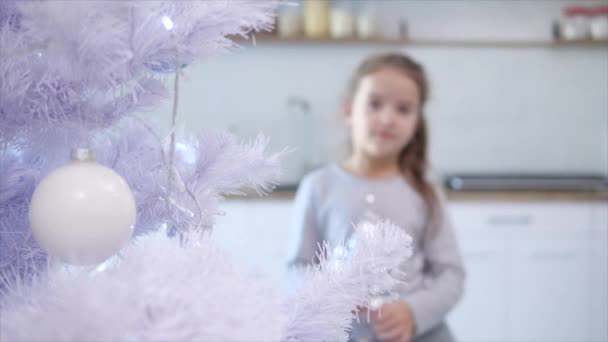 Mooi klein meisje komen naar de kerstboom op de voorgrond van wazig achtergrond en zet een glas schoonheid op een van takken. — Stockvideo