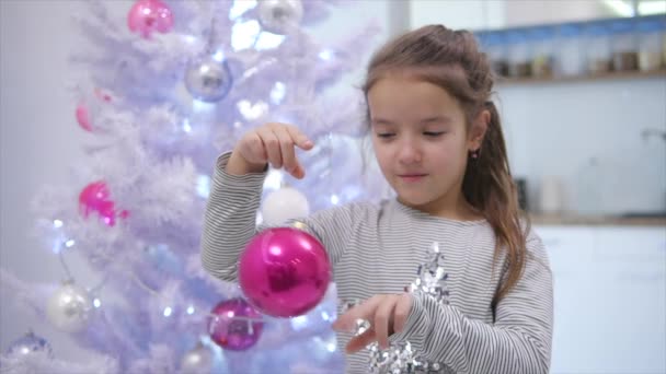 Χαρούμενο χαριτωμένο παιδί που παίζει με ένα ροζ μπιχλιμπίδι, το κουνάει από άκρη σε άκρη, στέκεται κοντά στο χριστουγεννιάτικο δέντρο, χαμογελώντας. — Αρχείο Βίντεο