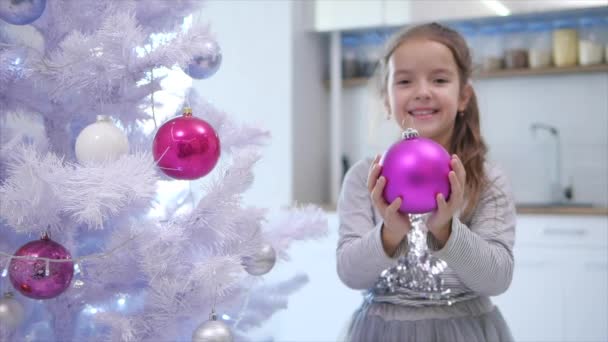 Mooi klein meisje is het uitbreiden van een roze bauble naar de camera, staan in de buurt van kerstboom, glimlachen. — Stockvideo